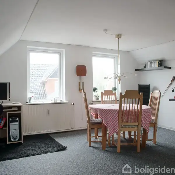 Et bord dækket med en rødternet dug står i midten af et rum omgivet af træstole. I hjørnet står et skrivebord med computer. Der er to vinduer med udsigt.