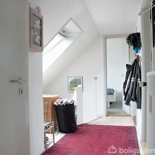 En aflang, hvid gang med åbent dørhul fører til et rum med seng. Sollys trænger ind gennem et skråvindue, mens en rød løber dækker gulvet. Vasketøjskurv står til venstre.