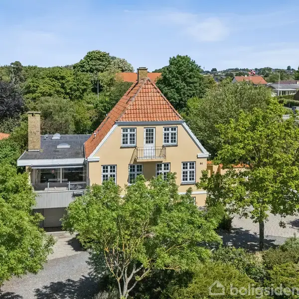 Et gult hus med rødt tegltag ligger omgivet af grønne træer i en villakvarter. Indkørsel med grus omgiver huset, der har en terrasse og flere vinduer.
