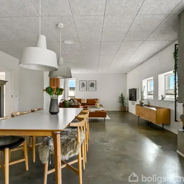 Spisebord med stole og lamper hænger over bordet. Pejsen er tændt og fjernsynet står modsat i stuen. Moderne, lys indretning med vinduer og sofa i baggrunden.