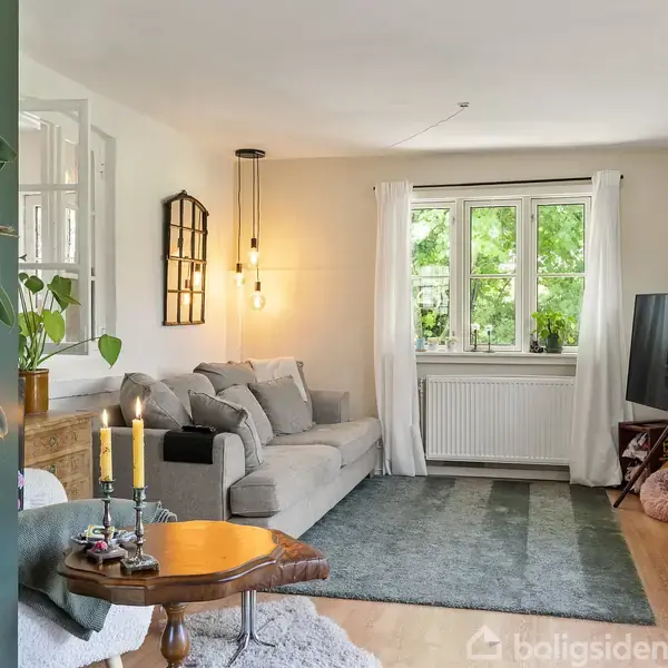 Sofa med puder står i en lys stue. En lampe hænger over sofaen. Foran sofaen er et kaffebord med lysestager. Store vinduer lukker lys og udsigt til bladtræer ind.