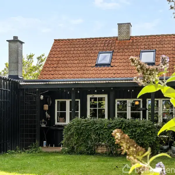 Et sort træhus med rødt tegltag og stor skorsten står i en have med grøn græsplæne. Vinduer på førstesalen har ovenlysvinduer, omgivet af buske og planter.