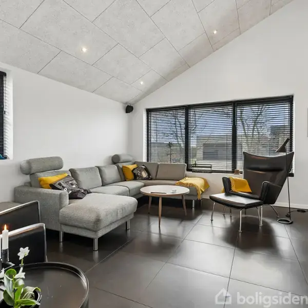 Grå hjørnesofa med gule puder står i en moderne dagligstue med skråloft. Store vinduer med persienner lukker lys ind. Skulpturel lænestol og små borde supplerer indretningen.