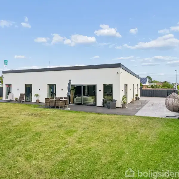 En moderne, hvid bygning med fladt tag ses med store vinduer mod en græsplæne. Foran huset er der en terrasse med møbler og en rund hængestol til venstre.
