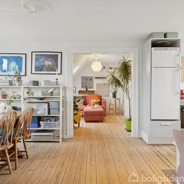 Spisebord og stole står på et trægulv, omgivet af lyse vægge med billeder. I baggrunden ses en stue med planter og sofa. Skydevindue til venstre og køkkenskabe til højre.