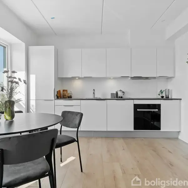 Et moderne køkken med hvide skabe og indbygget ovn. Foran ses et rundt spisebord med sorte stole. Sollys strømmer ind fra vinduet til venstre.