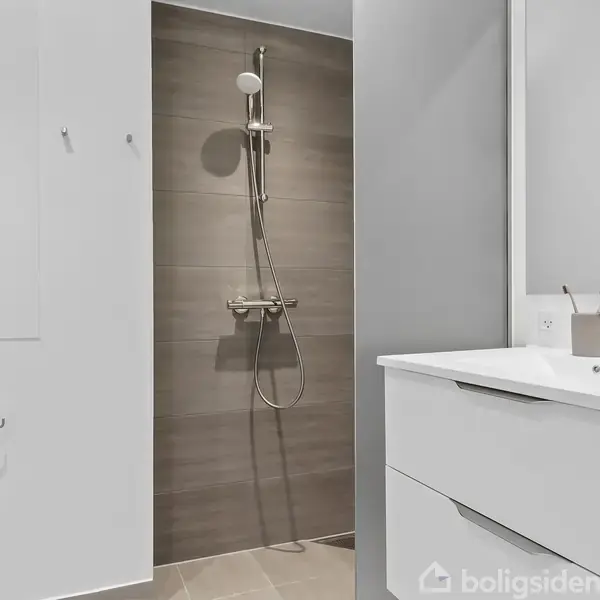Bruser hænger i et moderne badeværelse med grå fliser. Til højre er en hvid vask med tilbehør som sæbe og et spejl. Kontexten er minimalistisk indretning.