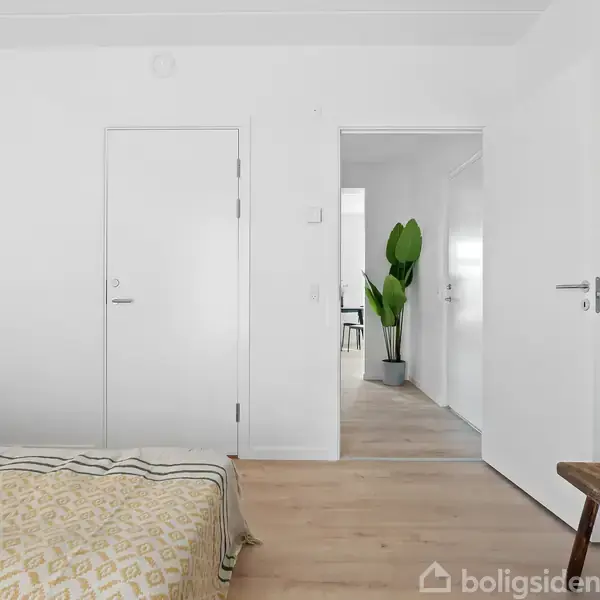 Seng står stille i et lyst soveværelse med en guldbelagte lampe på et bord. I baggrunden ses en åben dør, der leder til en gang med en grøn plante.