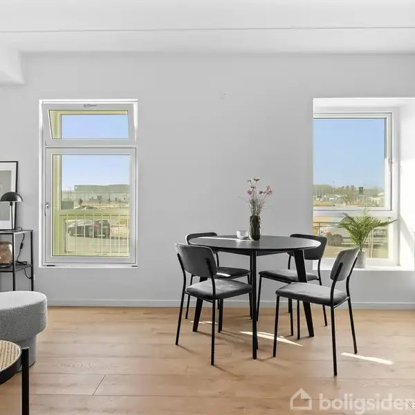 Et rundt spisebord med tre stole står i et moderne, lyst køkken-alrum med trægulv. To vinduer giver udsigt til landskabet udenfor. En vase med blomster pryder bordet.