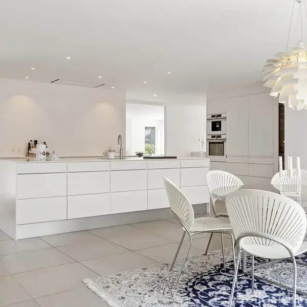Et moderne køkken- og spiseområde med hvide skabe og en ø øverst med apparater. Området har et rundt spisebord med hvide stole og en dekorativ, mønstret tæppe på gulvet. En kunstnerisk loftslampe hænger over bordet.