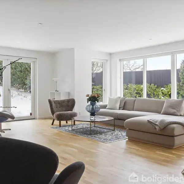 En lys stue med en stor hjørnesofa står mod vinduer, der lukker naturligt lys ind. Rummet er indrettet med en lænestol, sofabord, planter og trælaminatgulv.