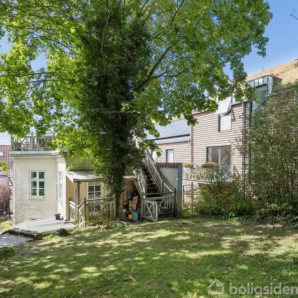 Et hvidt hus med en trappesektion ses midt i en have omgivet af store grønne træer, buske og en stensti. Baggrund med flere tilstødende bygninger.