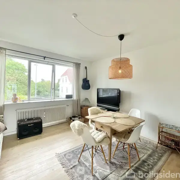 En spisestue indeholder et bord med fire stole i lys indretning. En lampe hænger over bordet, tv'et står ved væggen, og en guitar hænger ved vinduet med udsigt til træer.