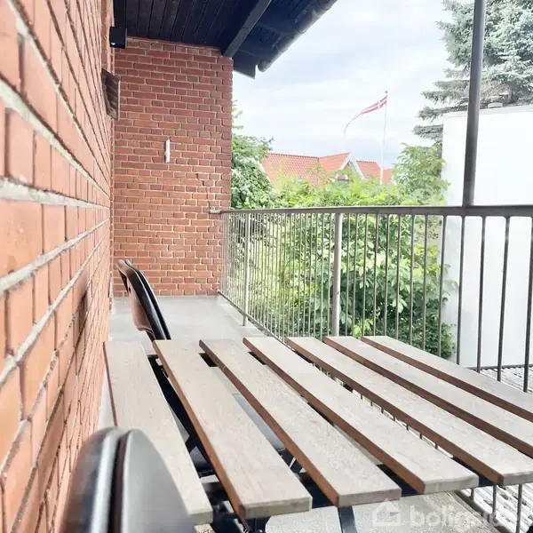 En balkon med en træbænk står ved siden af en rød murstensvæg. I baggrunden vajer et dansk flag over grønne træer og et rødt tag i et boligområde.