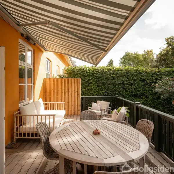 En rund udendørs træbord med flettede stole står på en terrasse. En sofa med puder er ved siden af, beskyttet af en stribemønstret markise. Omgivet af grønne buske.
