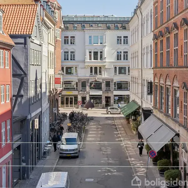 En gade set fra en høj bygning, hvor biler og cyklister bevæger sig mellem farverige, klassiske bygninger. Butikskilte og fortove pryder begge sider af vejen i bymiljøet.
