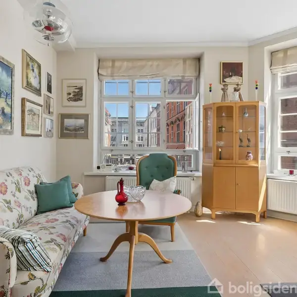 En stue med en blomsterprintet sofa står ved et rundt træbord med røde dekorationer. En grøn lænestol er ved vinduet, og en vitrineskab står til højre. Væggene er pyntet med malerier.