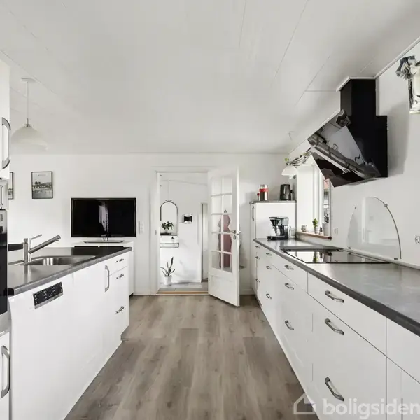 Et moderne køkken med hvide skabe og stålgreb; køkkenet er ryddeligt og velbelyst med et vindue, der slipper dagslys ind. En fransk dør åbner ud til et andet rum.