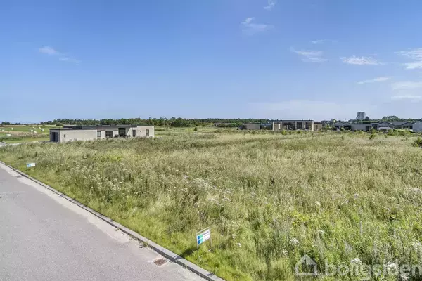 Grønt græsareal fyldt med ukrudt strækker sig mod enkelte lave bygninger i horisonten under en blå himmel. Skilt i forgrunden viser "Til salg," indikerer jord tilgængelig for køb.