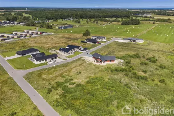 Flere huse står spredt i en udstykning omgivet af åbne græsarealer. Veje forbinder husene, og skove kan ses i baggrunden under en klar blå himmel.