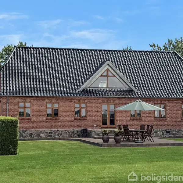 Et rødt murstenshus med sorte tagsten, terrasse, havebord og parasol. Placeret i en grøn have med træer i baggrunden.