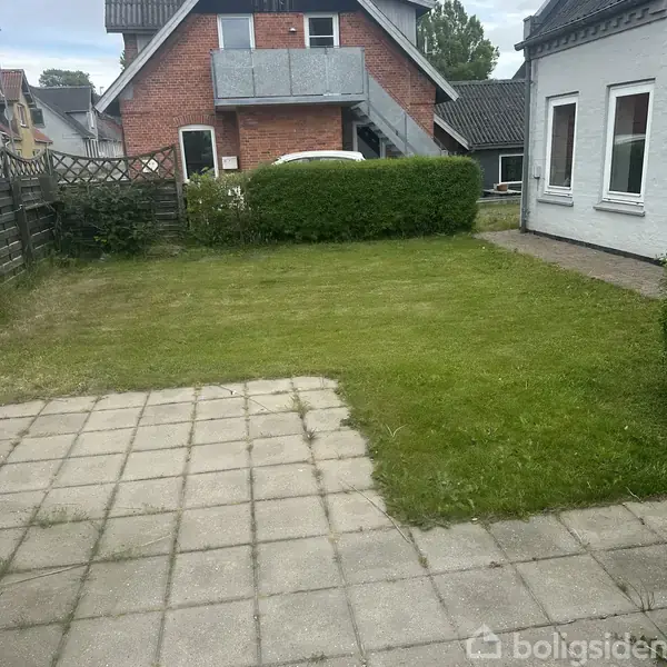 Græsplæne med fliseområde grænser op til et hvidt hus. Baggrunden viser et rødt murstenshus og velplejet hæk.