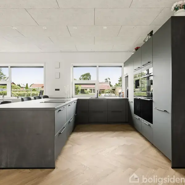 Køkken med grå indbyggede skabe og indbyggede hvidevarer. Detaljer inkluderer en køkkenø og spiseområde. Stort glasparti giver udsigt til have og naturligt lys.