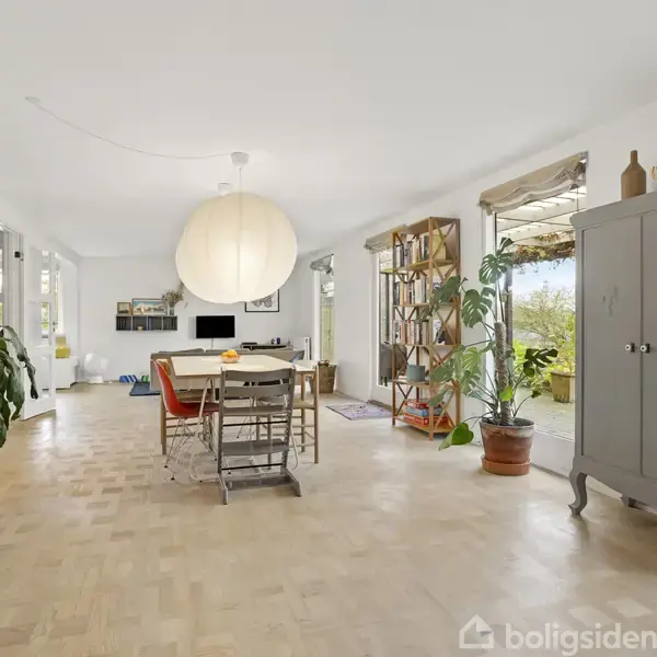 Stue med spisestue. Parquetgulv, spisebord med stole, stor rund lampe, gråt skab, reol med planter ved vinduer. Udenfor ses en terrasse. Vægdekorationer og naturlys tilstede.