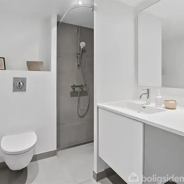 Toilet hænger på væggen; ved siden af er der en bruser i en aflukket sektion. En vask med underskab og spejl står til højre, dekoreret med blomster.