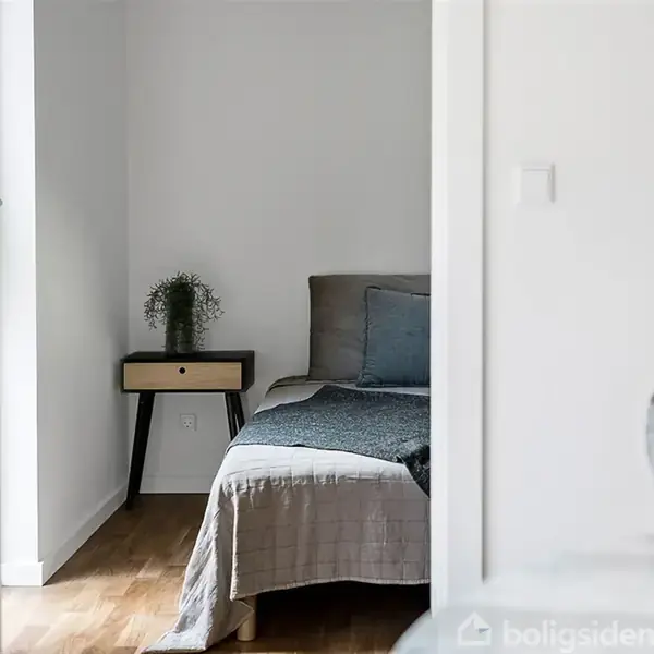 En seng med grå sengetæppe står i et hvidt værelse. Et lille natbord med en plante er ved siden af. Golvet er af træ.