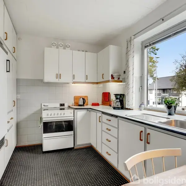 Et køkken med hvide skabe og sort bordplade. Komfur står ved siden af en elkedel og kaffemaskine. Gennem vinduet ses et træ udenfor. En stol er synlig i forgrunden.