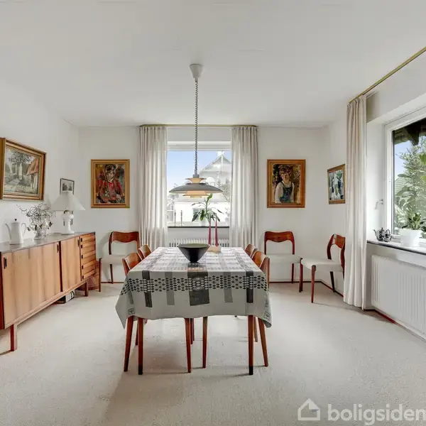 Spisebord med dug og skål står centralt, omgivet af træstole i en lys spisestue. Kommode med dekorationer og malerier på væggene. Vindue med udsigt i baggrunden.