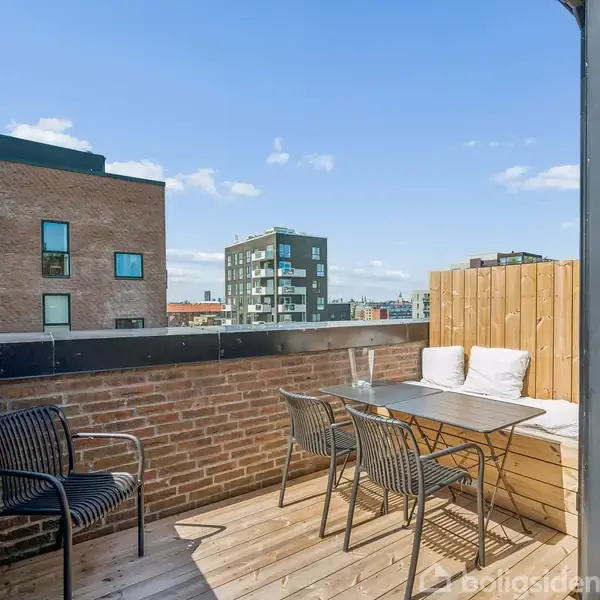 Lille tagterrasse med træbrætterrasse har et bord og to stole. Området er indhegnet af murstensvægge, med bybygninger i baggrunden under en blå himmel med få skyer.