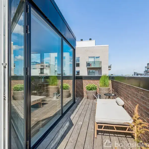 En træterrasse med en liggestol og planter står ved siden af store glaspartier. I baggrunden ses moderne lejlighedsbygninger under en klar blå himmel.