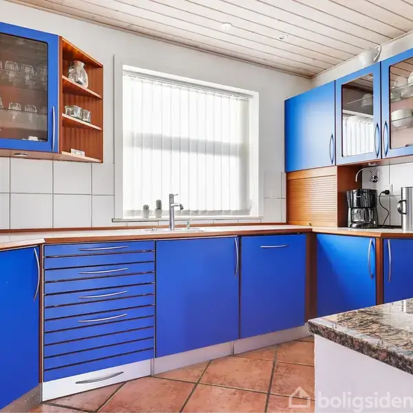 Køkkenskabe i blå farve omringer køkkenområdet, hvor en kaffemaskine står på en træbordplade. Fliser på væggen og en stor firkantet vindue giver naturligt lys.