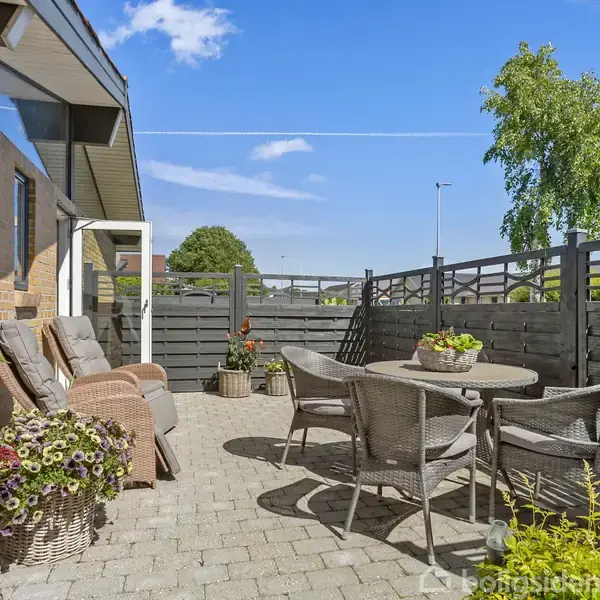 Terrasse med grå træhegn, rattanmøbler, og kurve med blomster. Solsenge står op ad murstensvæg. En blå himmel og træer ses i baggrunden, hvilket skaber en afslappende atmosfære.