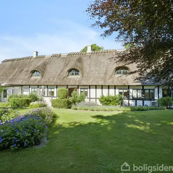 Bindingsværkshus med stråtag står stille. I forgrunden er der en velplejet græsplæne omgivet af blomsterbede. En glaspyramide til venstre tilføjer moderne elementer til den idylliske have.