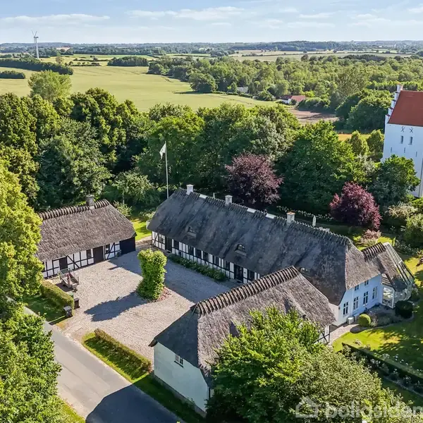 Bindingsværkshus med stråtag omkranset af frodige træer. En kirke med hvid facade og rødt tag ses i baggrunden. Landskabet er præget af grønne marker og skove.