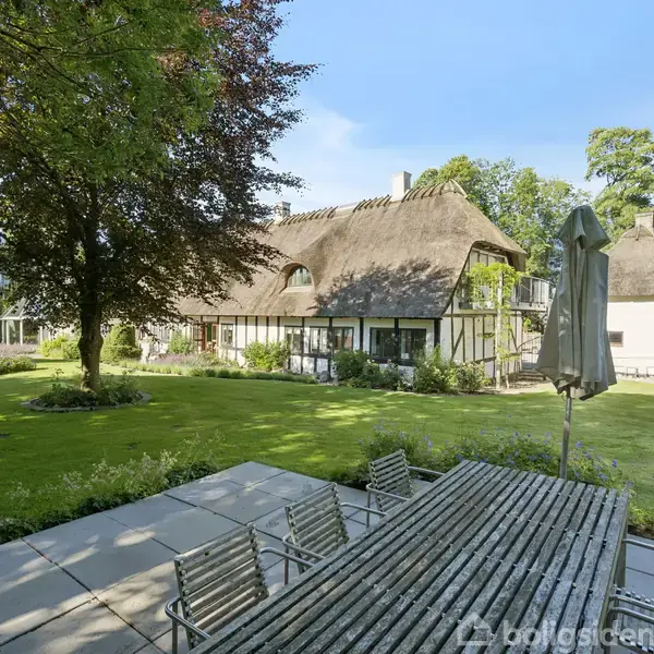 Et landhus med stråtag står i en frodig have. En stor træterrasse med bord og stole fører op til huset. Græsset er grønt, omgivet af træer og buske.