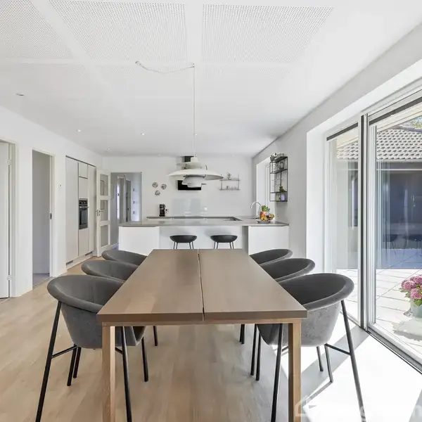 Et moderne køkken-alrum med et langt spisebord omgivet af seks stole. Der er store vinduer mod en terrasse og lysindfald. Køkkenet har hvide elementer og indbygget ovn.