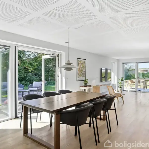 Spisestue med træbord og sorte stole står ved åbne terrassedøre. Dagslyset strømmer ind, bagved ses en terrasse med havemøbler og grøn have. Ingen tekst til stede.