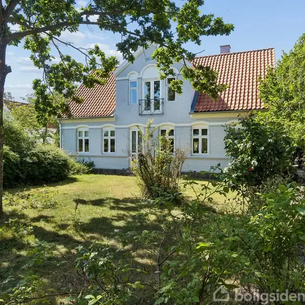 Et hvidt hus med rødt tegltag står stille i en grøn have med buske og træer.