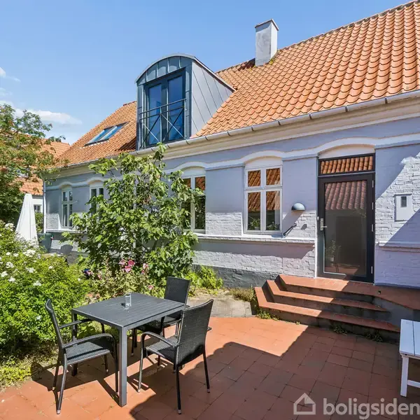 Hus med rødt tegltag og hvid facade, bord med stole står på en terrasse med røde fliser. Grønt buskads omgiver området under en blå himmel.