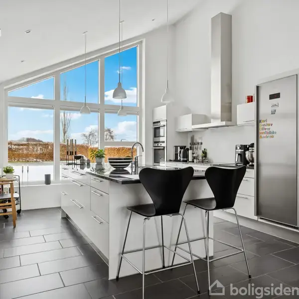 Køkken med moderne indretning; køkkenø med to sorte stole, stor stål-køleskab, og store vinduer viser sneklædt have. Lysegrå flisegulv og indbyggede loftslamper. Ingen tekst fundet.