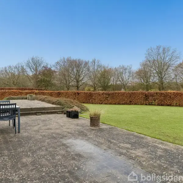 Terrassemøbler står på en flisebelagt terrasse ved et hus, omgivet af en grøn have med buske og træer i baggrunden under en blå himmel.