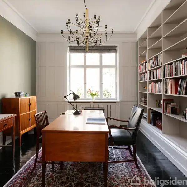 Et kontor med et træskrivebord og stol, oplyst af en vindueslampe, omgivet af bogreoler fyldt med bøger. En lysekrone hænger fra loftet. En tæppe pryder gulvet.