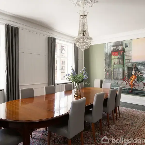 Et aflångt spisebord med otte polstrede stole står i en elegant spisestue. En stor maleri af en cyklist pryder den grønne væg. Store vinduer giver naturligt lys.