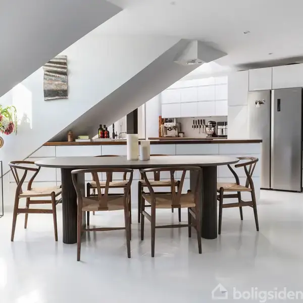 Et spisebord med seks stole står i et lyst, moderne køkken med skrå loftvægge. Ved siden af bordet er der et køleskab, planter, og en kommode med vaser og køkkenudstyr.