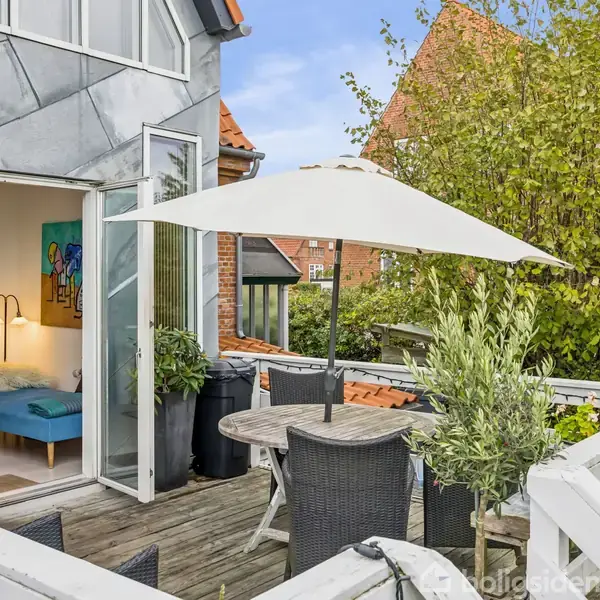 Et terrassebord med stole står under en parasol på en træterrasse. Åbne døre fører til et værelse med en seng. Omgivet af planter og træer.