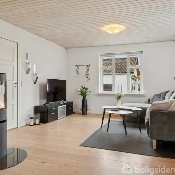 En moderne stue med en rund brændeovn til venstre, der brænder. En grå hjørnesofa står til højre omkring to små borde. Store vinduer giver naturligt lys.